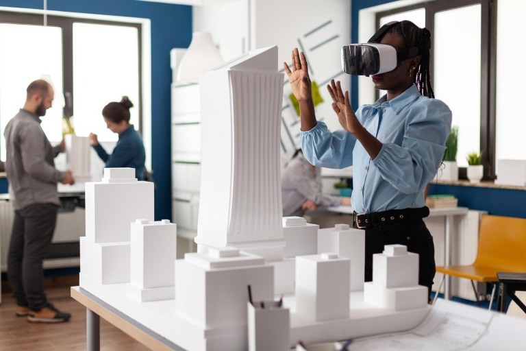 An image showing a woman wearing virtual reality goggles interacts with a model city made of white buildings. People work in the background of a modern office