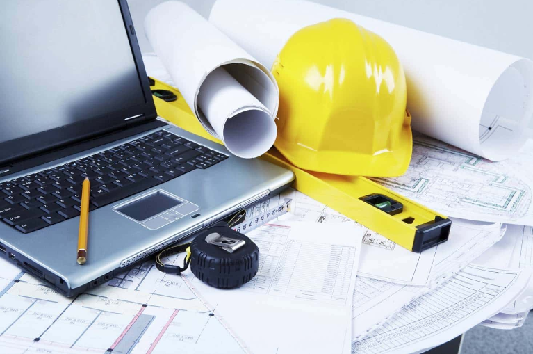 A laptop, construction plans, and a helmet on a desk