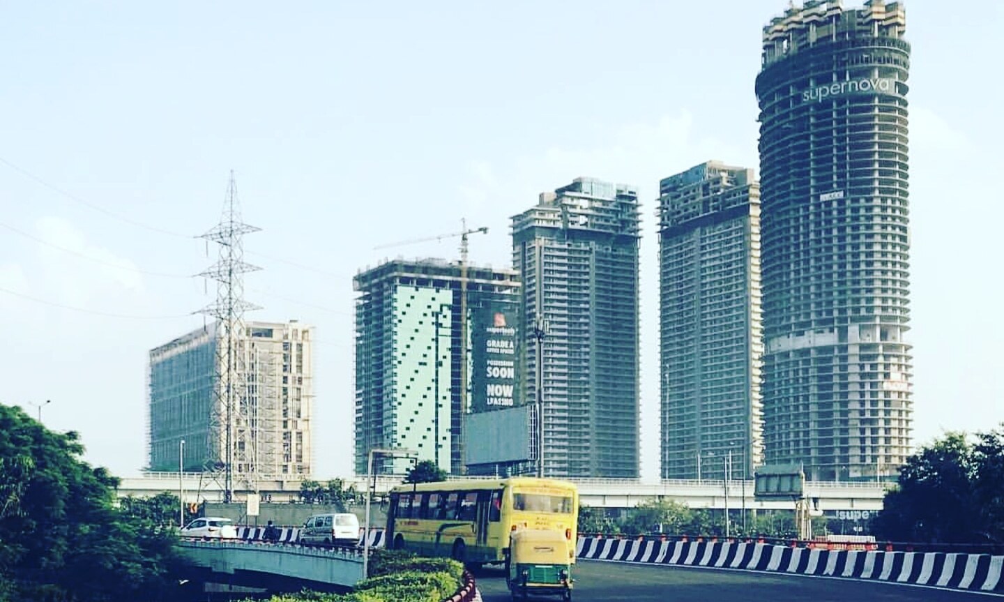 Supernova Spira skyscraper rising above the Noida city skyline, surrounded by other high-rise buildings under a clear blue sky.