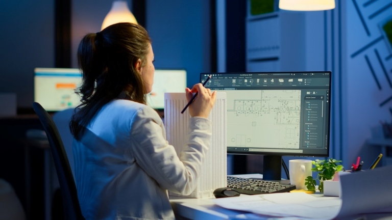 The image shows an engineer working with CAD, displaying a 3D model and digital blueprints on screens while comparing them to a physical blueprint.
