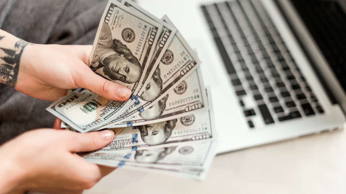 A person holding cash in their hand beside a laptop